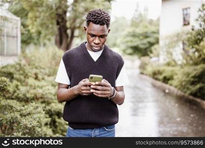 Young sad African American man in bomber jacket in park or street outdoor. Fall or spring season. Young sad African man typing message, texting on phone in rain outdoor