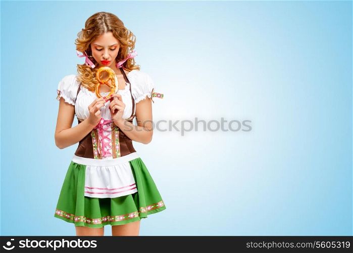 Young sad sexy Oktoberfest woman wearing a traditional Bavarian dress dirndl holding a pretzel in hands on blue background.