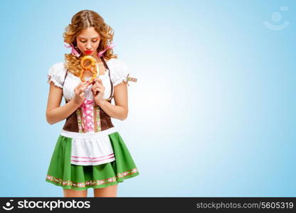 Young sad sexy Oktoberfest woman wearing a traditional Bavarian dress dirndl holding a pretzel in hands on blue background.