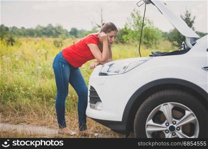 Young sad woman looking at engine of broken car