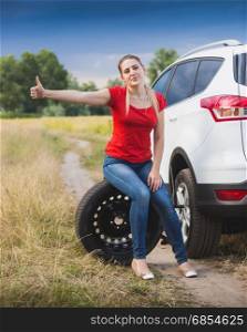 Young sad woman sitting on wheel at broken car and hitchhiking