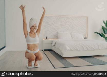 Young satisfied beautiful lady in perfect shape squatted on scales in white underwear and towel wrapped on head after morning shower in bedroom, throwing her hands up celebrating her weight loss. Young satisfied beautiful lady in perfect shape squatted on scales