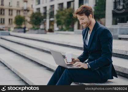 Young satisfied businessman in stylish dark blue suit sitting on stairs near office center outside with laptop and coffee to go in sunny summer day, working remotely online while having lunch break. Young satisfied businessman in stylish suit sitting on stairs outside with laptop and coffee to go