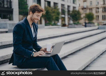 Young satisfied businessman in stylish dark blue suit sitting on stairs near office center outside with laptop and coffee to go in sunny summer day, working remotely online while having lunch break. Young satisfied businessman in stylish suit sitting on stairs outside with laptop and coffee to go