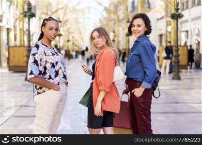 Young satisfied diverse ladies in casual clothes with paper bags looking at camera while standing on blurred street. Multiracial female friends standing on city street with purchases