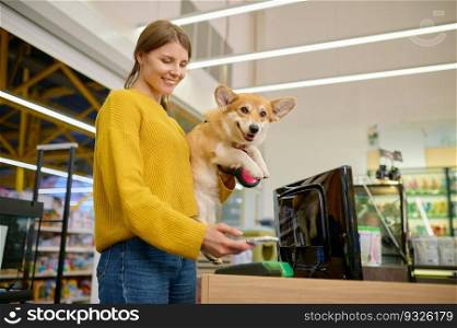 Young satisfied woman holding her corgi dog on hands paying for purchase at pet shop counter desk. Female customer using mobile app for wireless payment. Young satisfied woman paying for purchase at pet shop counter desk