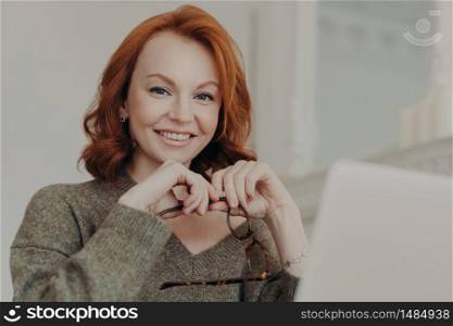 Young satisfied woman with foxy hair works freelance on modern laptop computer, uses wireless internet for publication, searches information, holds glasses, wears warm sweater, smiles happily