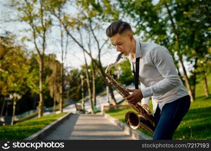 Young saxophonist plays melody on golden saxophone in summer park. Musician with sax outdoors, musical performance in nature, jazz performer. Young saxophonist plays melody on golden saxophone