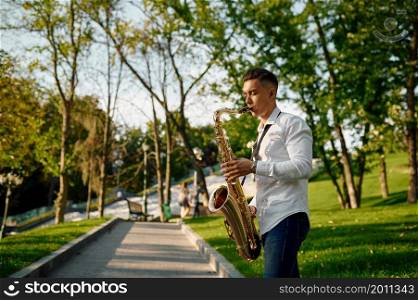 Young saxophonist plays the saxophone in summer park. Musician with sax outdoors, musical performance in nature, jazz melody performer. Young saxophonist plays the saxophone in park