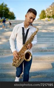 Young saxophonist plays the saxophone on stairs in summer park. Musician with sax outdoors, musical performance in nature, jazz melody performer. Saxophonist plays the saxophone on stairs in park