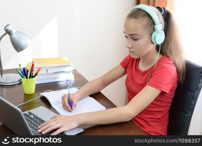 Young schoolgirl with headphones is doing a homework or learning foreign language on the laptop in her room.