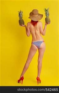 Young semi-naked girl in retro striped bathing shorts and high-heeled shoes and a wide-brimmed hat holds two pineapples in her hands while standing with her back to the viewer on a bright yellow background. girl With Pineapple