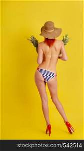 Young semi-naked girl in retro striped bathing shorts and high-heeled shoes and a wide-brimmed hat holds two pineapples in her hands while standing with her back to the viewer on a bright yellow background. girl With Pineapple