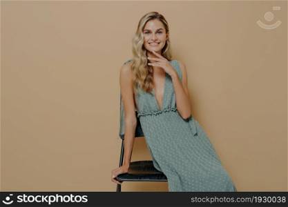Young sensual and flirty blonde woman posing in elegant romantic green dress, leaning on black chair against beige studio wall and looking at camera with delight and joy, touching her chin gently. Young sensual and flirty blonde woman posing in elegant romantic green dress in studio