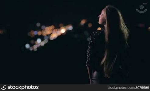 Young sensual woman is standing on the observation deck and enjoying panorama of city at night. The wind is blowing in her long nice hair. On background city streetlights.