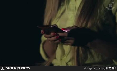 Young sensual woman texting on smartphone outdoors at night on blurred night city lights background