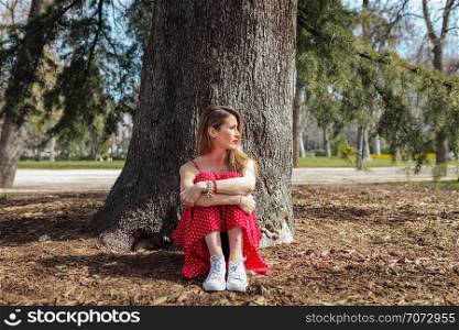 Young serious blond woman siting near tree with red long dress