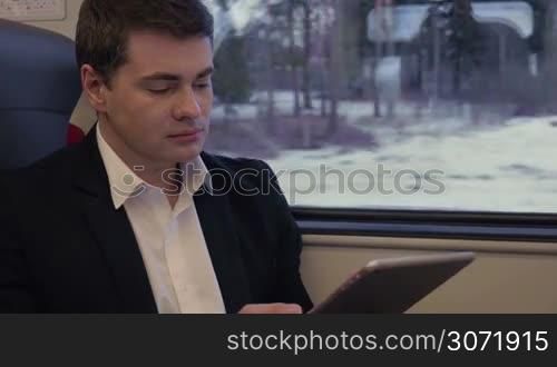 Young serious businessman traveling by train and using tablet computer, sometimes he getting distracted with view in the window. Business trip
