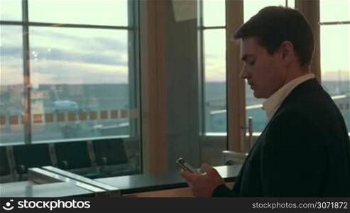 Young serious businessman typing text message on smart phone and looking out the window at airport