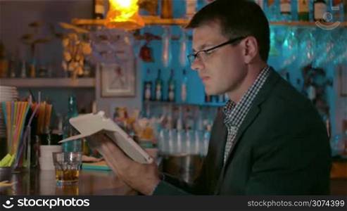 Young serious businessman with tablet in hands is sitting in the bar and drinking something from the glass.