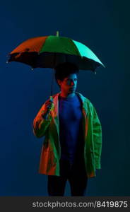 Young serious man wearing raincoat standing under umbrella over dark studio background under neon light illumination. Man in raincoat under umbrella over dark studio background