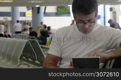 Young serious man with digital tablet in waiting-room at the airport or station. He using pad to work or pass the time before departure