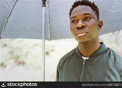 Young serious or sad African American man with umbrella, under rain on street outdoor. Fall or spring season. Young African American man under black umbrella in rain, sad. Fall or spring weather