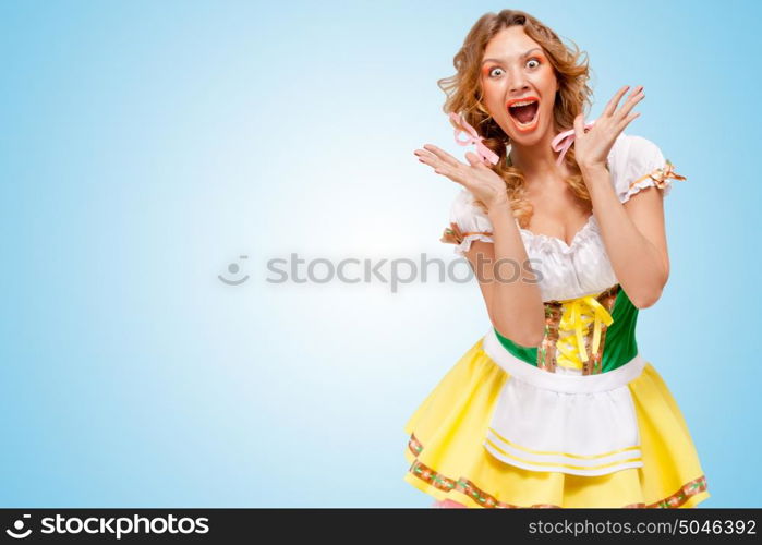 Young sexy astonished Oktoberfest woman wearing a traditional Bavarian dress dirndl, showing happy emotions on blue background.