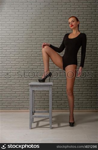 young sexy girl with wet red hair in a black sports bodysuit and mesh pantyhose on a rough wooden stool against a gray brick wall. girl with stool