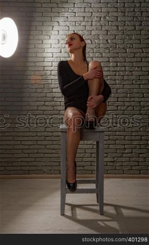 young sexy girl with wet red hair in a black sports bodysuit and mesh pantyhose on a rough wooden stool against a gray brick wall. girl with stool