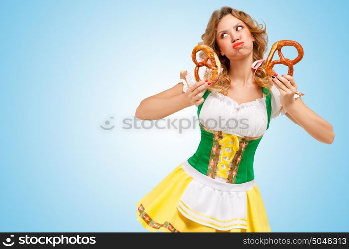 Young sexy offended Oktoberfest woman wearing a traditional Bavarian dress dirndl holding two pretzels and puffing her cheeks on blue background.