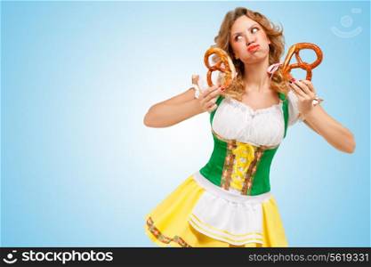 Young sexy offended Oktoberfest woman wearing a traditional Bavarian dress dirndl holding two pretzels and puffing her cheeks on blue background.