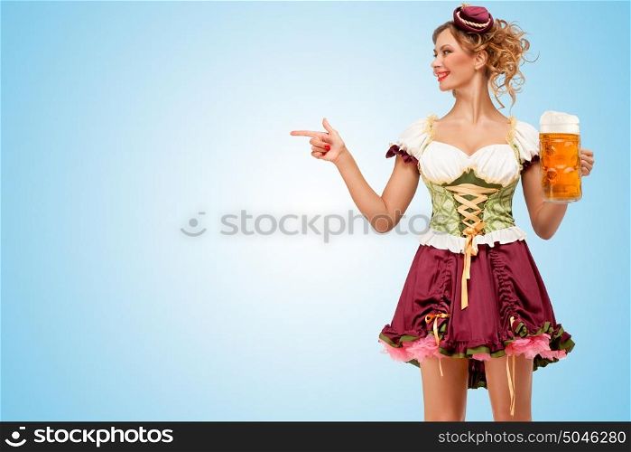 Young sexy Oktoberfest waitress wearing a traditional Bavarian dress dirndl holding a beer mug, and pointing aside on blue background.