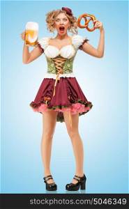Young sexy Oktoberfest waitress wearing a traditional Bavarian dress dirndl holding a pretzel and beer mug, and laughing happily on blue background.