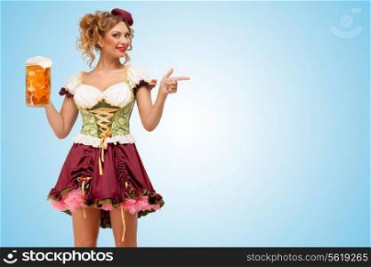Young sexy Oktoberfest waitress wearing a traditional Bavarian dress dirndl holding a beer mug, and pointing aside on blue background.