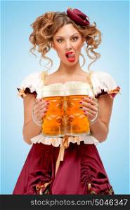 Young sexy Oktoberfest waitress, wearing a traditional Bavarian dress dirndl, serving two big beer mugs in a tavern and licking her lips on blue background.