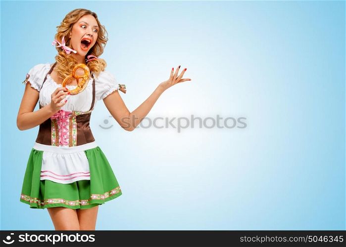Young sexy Oktoberfest woman wearing a traditional Bavarian dress dirndl dancing with a pretzel in hands on blue background.