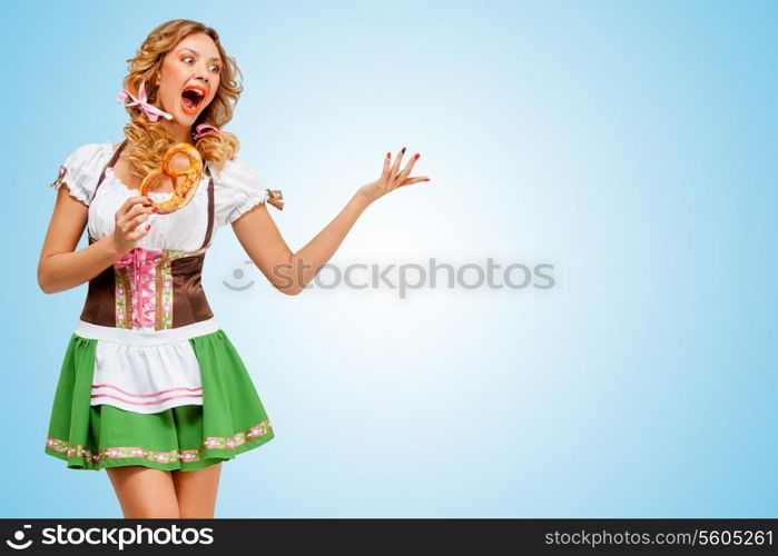 Young sexy Oktoberfest woman wearing a traditional Bavarian dress dirndl dancing with a pretzel in hands on blue background.