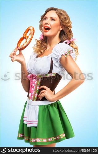 Young sexy Oktoberfest woman wearing a traditional Bavarian dress dirndl eating a pretzel on blue background.