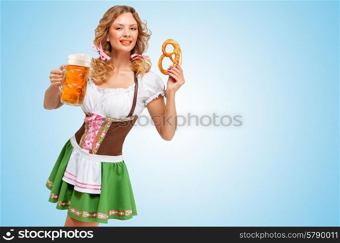 Young sexy Oktoberfest woman wearing a traditional Bavarian dress dirndl holding a pretzel and beer mug in hands on blue background.