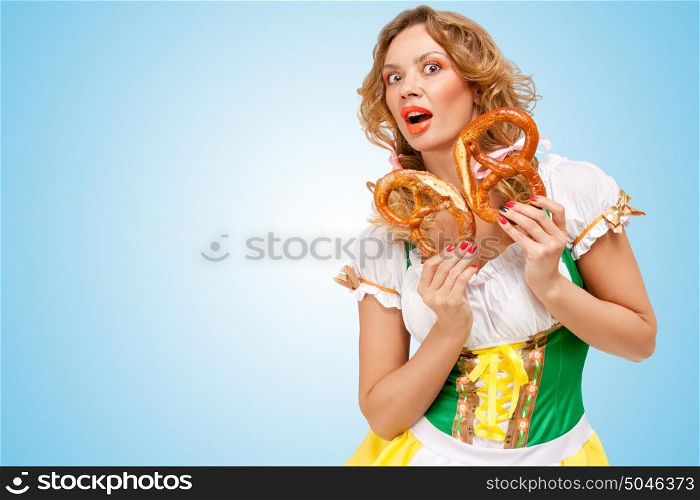 Young sexy Oktoberfest woman wearing a traditional Bavarian dress dirndl holding two pretzels and feeling scared on blue background.