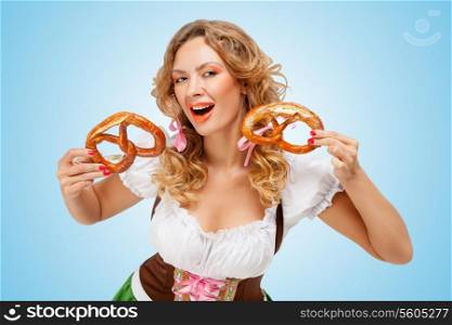 Young sexy Oktoberfest woman wearing a traditional Bavarian dress dirndl laughing happily and holding two pretzels in hands on blue background.