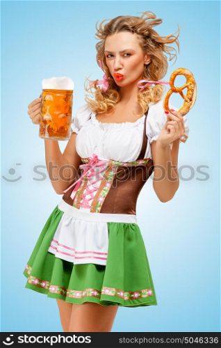Young sexy Oktoberfest woman wearing a traditional Bavarian dress dirndl posing with a pretzel and beer mug in hands on blue background.