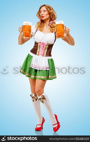 Young sexy Oktoberfest woman wearing a traditional Bavarian dress dirndl serving two beer mugs on blue background.