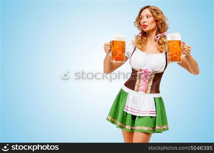 Young sexy Oktoberfest woman wearing a traditional Bavarian dress dirndl serving two beer mugs on blue background.