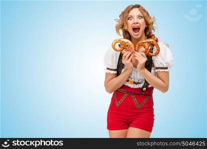 Young sexy Swiss woman wearing red jumper shorts with suspenders in a form of a traditional dirndl, holding with hunger two pretzels on blue background.