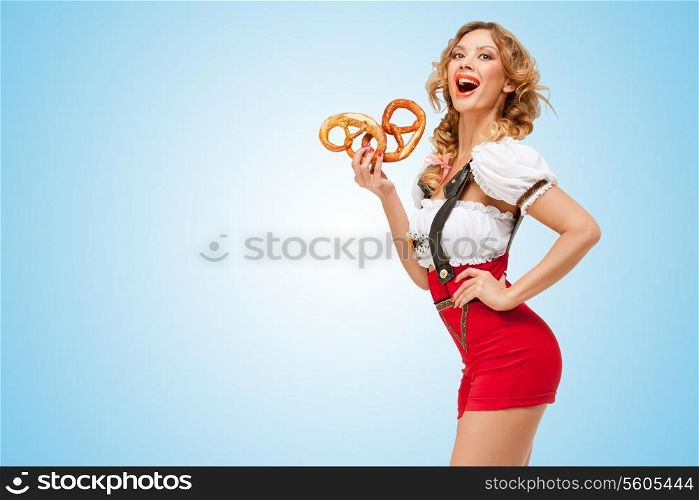 Young sexy Swiss woman wearing red jumper shorts with suspenders in a form of a traditional dirndl, eating two pretzels in her hand on blue background.