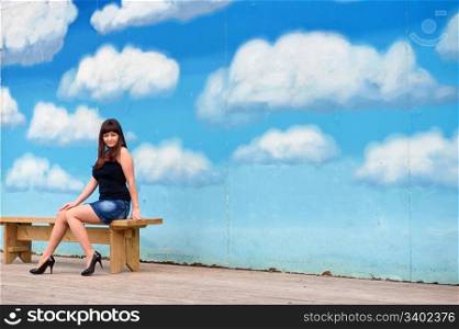 Young, sexy woman in short skirt sit on the bench on the background of blue sky drawing.