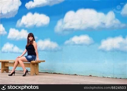 Young, sexy woman in short skirt sit on the bench on the background of blue sky drawing. 
