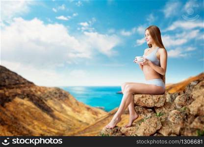 Young sexy woman sitting on the mountain with cup of coffee, good morning, ocean on background